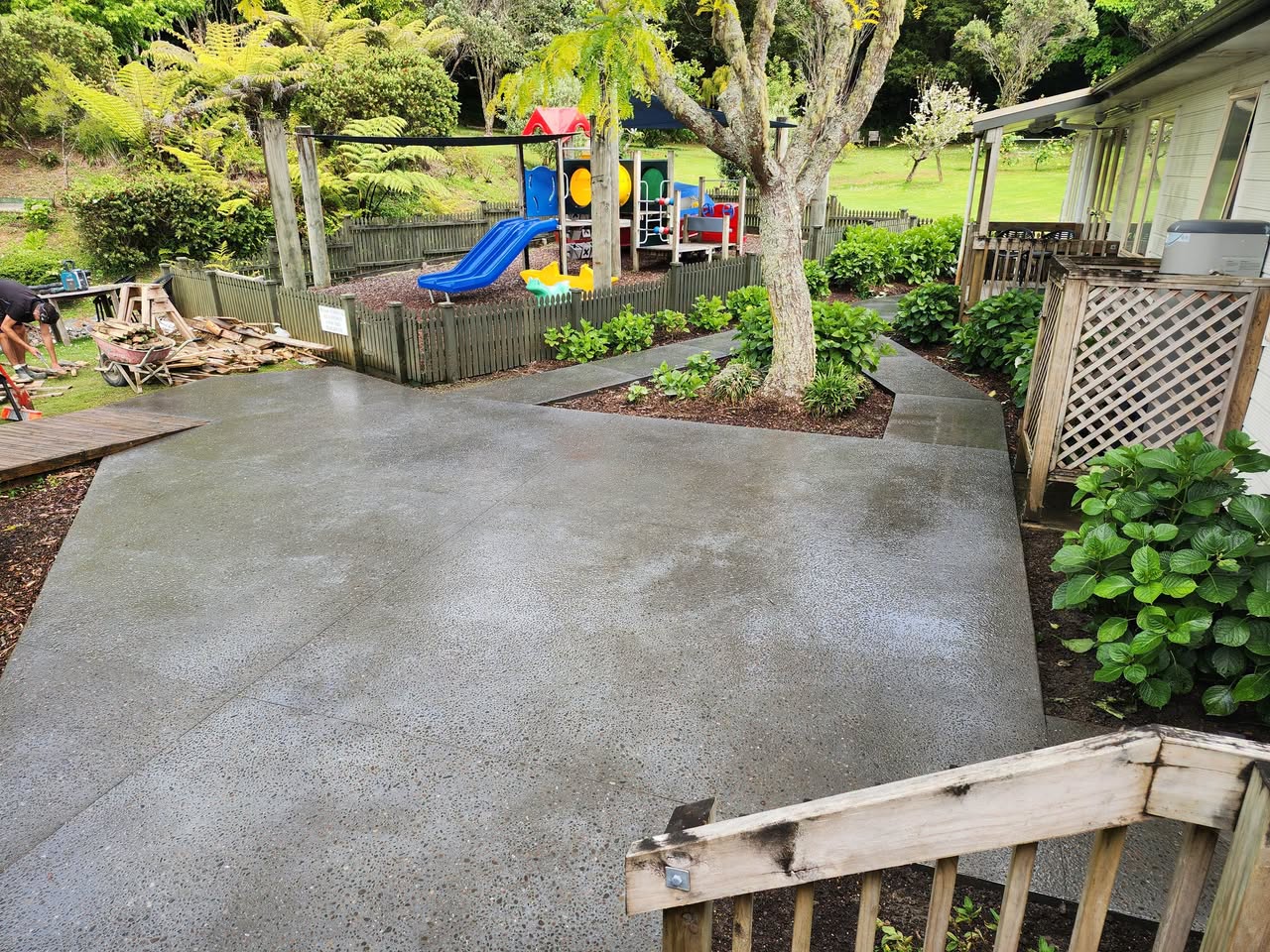 A large finished grey concrete area beside garden planting and play equipment.